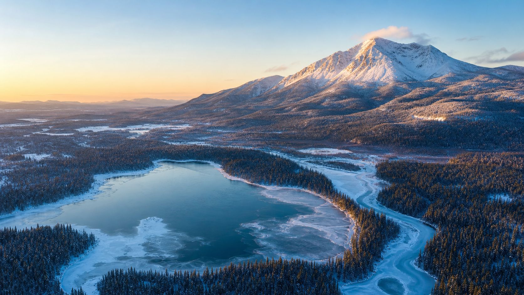 Winter aerial of a frozen Maine lake ringed by spruce forest below a snow-covered mountain at dawn